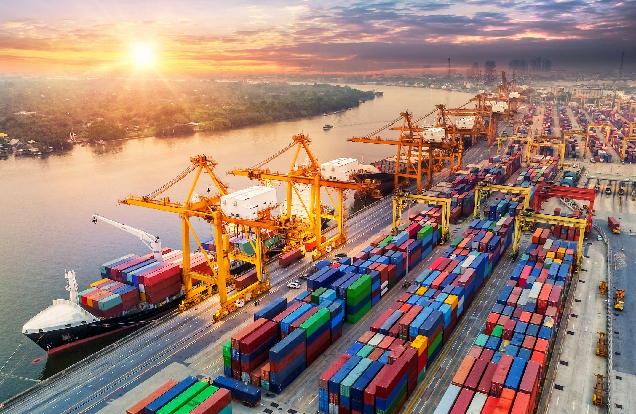 Container port with cargo ship, gantry cranes and stacked shipping containers at sunrise.